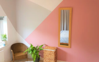 bedroom in geometric pattern painted on wall, grey cream and pink. Also contains a house plant, chair and laundry basket